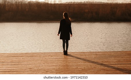 Full Body Back View Of Unrecognizable Female In Black Coat Standing Alone On Wooden Pier Near Calm River And Contemplating Sunset In Autumn Evening