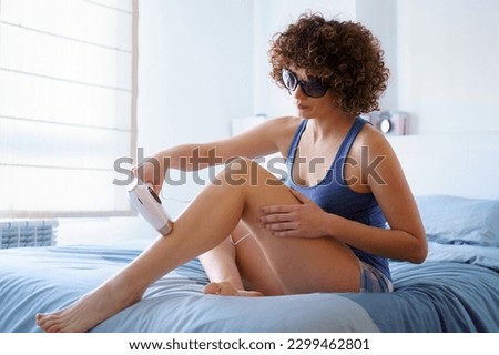 Full body of calm barefoot young female, in sleepwear and protective goggles with curly hairstyle sitting on comfortable bed and removing leg hair with pulsed light laser in bedroom