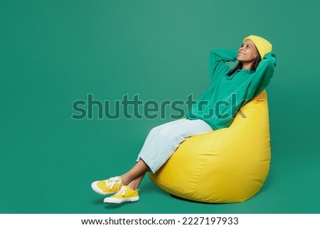 Full body calm little kid teen girl of African American ethnicity 13-14 year old wear casual hoody hat sit in bag chair hold hands behind neck isolated on plain dark green background Childhood concept