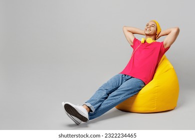 Full body calm relaxed young man of African American ethnicity 20s he wear pink t-shirt yellow hat headphones sit in bag chair hold hands behind neck take rest isolated on plain grey background studio - Powered by Shutterstock