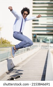 Full Body Of Carefree African American Male In Smart Casual Style Jumping Above Skateboard In City And Looking At Camera