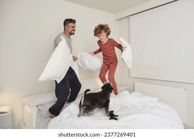 Full body of cheerful adult father and curly haired son in pajamas jumping on bed with pillows while playing with dog - Powered by Shutterstock