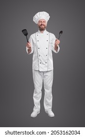 Full Body Of Cheerful Bearded Male Chef In White Uniform And Hat Showing Spatula And Spoon While Preparing Food In Restaurant Kitchen, Isolated On Gray Background