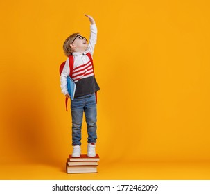 Full Body Cheerful Boy In Glasses Standing On Stack Of Textbooks And Raising Arm While Trying To Be Higher During School Studies Against Yellow Background
