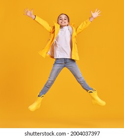 Full body of cheerful energetic preteen girl in yellow raincoat and gumboots leaping with outstretched arms and looking at camera against yellow background
