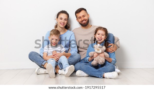 Full body of cheerful family with little kids\
looking at camera while sitting together on floor near wall in\
empty room of new home