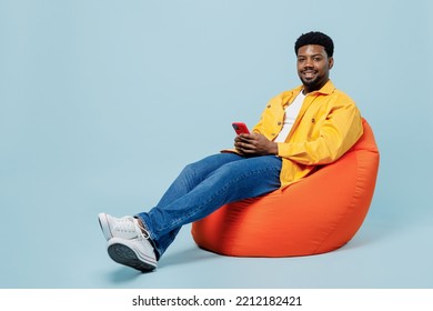 Full Body Cheerful Happy Young Man Of African American Ethnicity 20s In Yellow Shirt Sit In Bag Chair Hold In Hand Use Mobile Cell Phone Isolated On Plain Pastel Light Blue Background Studio Portrait.