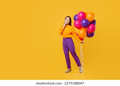 Full body cheerful side profile view happy fun young woman wearing casual clothes celebrating holding face bunch of balloons isolated on plain yellow background. Birthday 8 14 holiday party concept