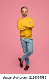 Full body of cheerful stylish male hipster in bright clothes standing with crossed legs and arms on pink background in studio