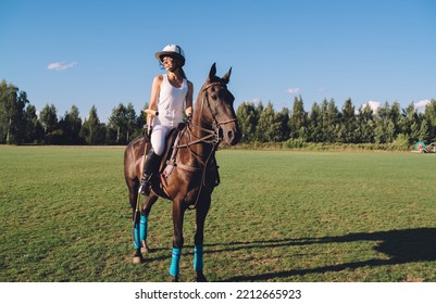 Full Body Of Cheerful Young Female Equestrian In Helmet And Boots With Riding Crop Sitting On Saddle On Brown Horse During Training At Field While Looking Away