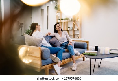 Full body of cheerful young women sitting with crossed legs on comfortable couch in stylish living room and talking to each other - Powered by Shutterstock