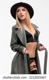 Full Body Of Confident Young Alluring Female Model With Long Blond Hair In Sexy Lingerie High Heeled Leather Boots Coat And Hat Standing Against White Background With Hand On Waist