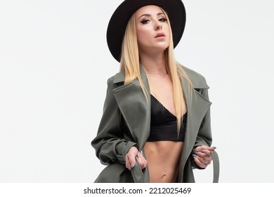 Full Body Of Confident Young Alluring Female Model With Long Blond Hair In Sexy Lingerie High Heeled Leather Boots Coat And Hat Standing Against White Background With Hand On Waist