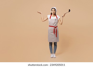 Full body confused sad shocked young housewife housekeeper chef baker latin woman wear apron toque hat hold in hand laddle spread hand isolated on plain pastel light beige background Cook food concept