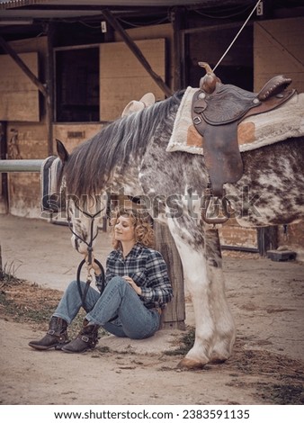 Full body of content horsewoman sitting at wooden post with spotted horse with bridle and saddle in countryside on summer day