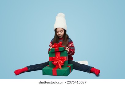 Full body of cute little girl in knitted sweater and hat sitting on stack of gift boxes and opening christmas presents against blue background - Powered by Shutterstock