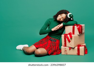 Full body cute merry little kid teen girl wear hat casual clothes sit put hand on present boxes with gift ribbon bow posing isolated on plain green background. Happy New Year Christmas holiday concept - Shutterstock ID 2396398837