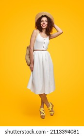 Full body of delighted curly haired female wearing straw hat and white sundress standing on yellow background in studio and looking away