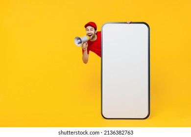 Full body delivery guy employee man in red cap T-shirt uniform workwear work as dealer courier stand behind mobile phone blank screen workspace shout in megaphone isolated on plain yellow background