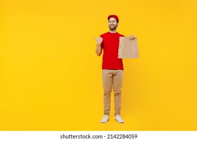 Full Body Delivery Guy Employee Man In Red Cap T-shirt Uniform Workwear Work As Dealer Courier Hold Brown Blank Craft Paper Takeaway Bag Mock Up Point Finger Aside Isolated On Plain Yellow Background