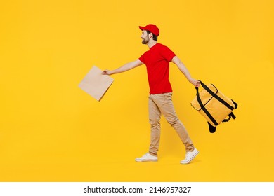 Full body delivery guy employee man in red cap T-shirt uniform work as dealer courier hold brown blank craft paper takeaway bag mock up thermal food bag backpack go isolated on plain yellow background
