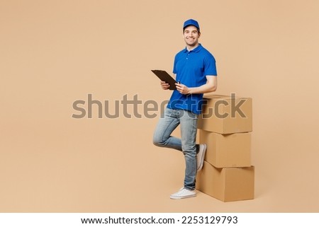 Full body delivery guy employee man wears blue cap t-shirt uniform workwear work as dealer courier stand near stack cardboard boxes hold clipboard papers documents isolated on plain beige background Stockfoto © 