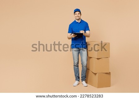 Full body delivery guy employee man wear blue cap t-shirt uniform workwear work as dealer courier stand near stack cardboard boxes hold clipboard with paper document isolated on plain beige background