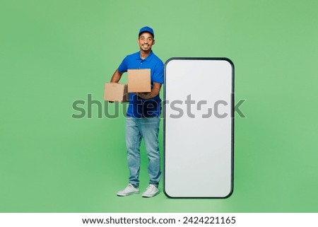 Full body delivery guy employee man wears blue cap t-shirt uniform workwear work as dealer courier big huge blank screen mobile cell phone hold box isolated on plain green background. Service concept