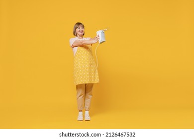 Full Body Elderly Happy Cheerful Satisfied Housekeeper Housewife Woman 50s In Orange Apron Holding Mixer Cooking Isolated Plain On Yellow Background Studio Portrait. People Household Lifestyle Concept