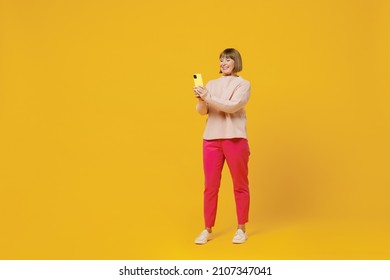 Full Body Elderly Happy Woman 50s Wears Pink Casual Knitted Sweater Using Mobile Cell Phone Chatting Browsing Internet Isolated On Plain Yellow Background Studio Portrait. People Lifestyle Concept.