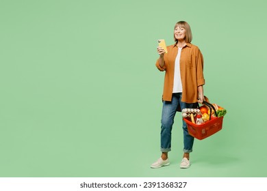 Full body elderly happy woman wear brown shirt casual clothes hold shopping paper bag with food products use mobile cell phone read list isolated on plain green background. Delivery service from shop - Powered by Shutterstock