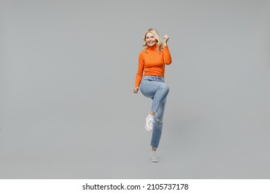 Full Body Elderly Overjoyed Excited Happy Blonde Woman 50s In Orange Turtleneck Do Winner Gesture Raise Up Leg Clench Fist Isolated On Plain Grey Background Studio Portrait. People Lifestyle Concept