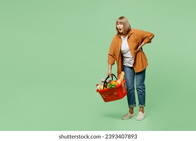 Full body elderly sad sick ill woman wear brown shirt casual clothes hold back waist carry heavy bag with food products after shopping isolated on plain green background. Delivery service from shop - Powered by Shutterstock