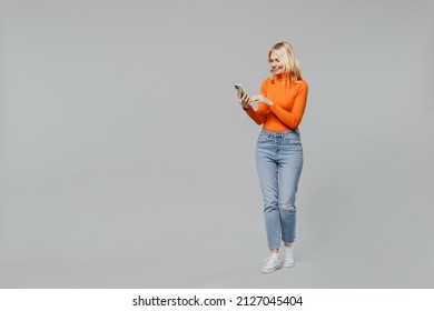 Full body elderly satisfied smiling happy blonde woman 50s in orange turtleneck hold in hand use mobile cell phone chatting browsing internet isolated on plain grey color background studio portrait.