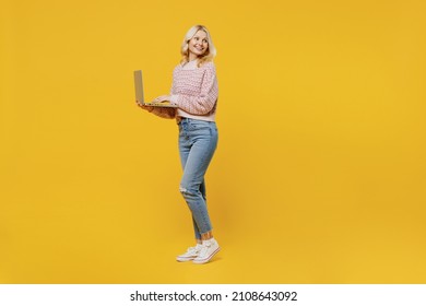 Full Body Elderly Smiling Blonde Caucasian Woman 50s In Pink Sweater Hold Use Work On Laptop Pc Computer Look Aside Isolated On Plain Yellow Color Background Studio Portrait. People Lifestyle Concept