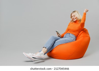 Full body elderly smiling blonde woman 50s in orange turtleneck sit in bag chair hold in hand use mobile cell phone do winner gesture isolated on plain grey background studio People lifestyle concept - Shutterstock ID 2127045446