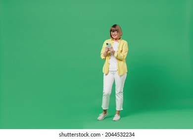 Full Body Elderly Smiling Cool Happy Woman 50s In Glasses Yellow Shirt Using Hold In Hand Mobile Cell Phone Browse Internet Isolated On Plain Green Background Studio Portrait People Lifestyle Concept