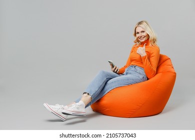 Full Body Elderly Smiling Fun Blonde Woman 50s In Orange Turtleneck Sit In Bag Chair Hold In Hand Use Mobile Cell Phone Show Thumb Up Isolated On Plain Grey Background Studio People Lifestyle Concept