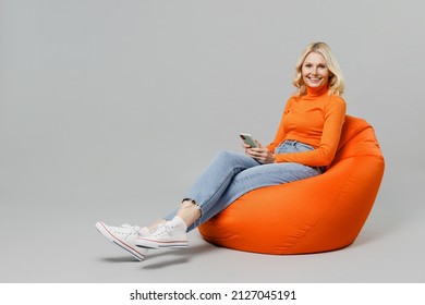 Full Body Elderly Smiling Happy Blonde Woman 50s In Orange Turtleneck Sit In Bag Chair Hold In Hand Use Mobile Cell Phone Isolated On Plain Grey Background Studio Portrait. People Lifestyle Concept