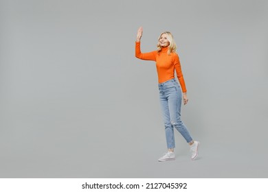 Full Body Elderly Smiling Happy Blonde Caucasian Friendly Cool Woman 50s In Orange Turtleneck Walking Going Waving Hand Isolated On Plain Grey Color Background Studio Portrait People Lifestyle Concept