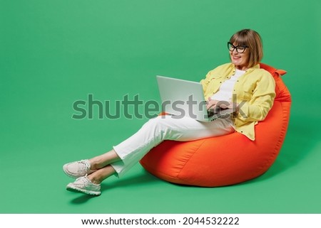 Full body elderly smiling happy woman 50s in glasses yellow shirt sit in bag chair resting relax hold use work on laptop pc computer isolated on plain green background studio People lifestyle concept Foto stock © 