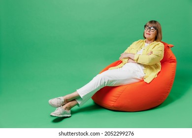 Full body elderly smiling minded happy woman 50s in glasses yellow shirt sit in bag chair resting relax during leisure time isolated on plain green background studio portrait People lifestyle concept
