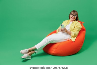 Full Body Elderly Smiling Satisfied Happy Woman 50s In Glasses Yellow Shirt Sit In Bag Chair Resting Relax Hold Use Mobile Cell Phone Isolated On Plain Green Background Studio People Lifestyle Concept