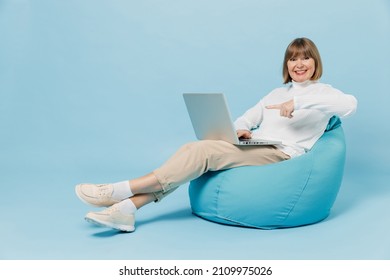 Full Body Elderly Smiling Woman 50s In White Knitted Sweater Sit In Bag Chair Hold Use Work Point On Laptop Pc Computer Isolated On Plain Blue Color Background Studio Portrait People Lifestyle Concept