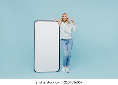 Full body elderly woman 50s in shirt near big white empty blank poster billboard for promotional content, place for text or image point finger up with new idea isolated on plain pastel blue background - Shutterstock ID 2154836069
