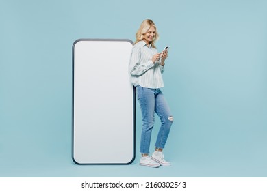 Full body elderly woman 50s in striped shirt near big white blank poster billboard for promotional content, place for text or image use mobile cell phone isolated on plain pastel light blue background - Shutterstock ID 2160302543