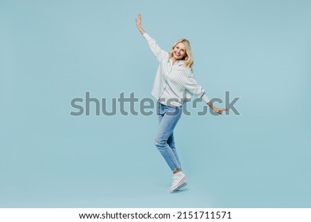 Full body elderly woman 50s wear casual striped shirt looking camera lean back stand on toes dance fooling around isolated on plain pastel light blue color background studio People lifestyle concept