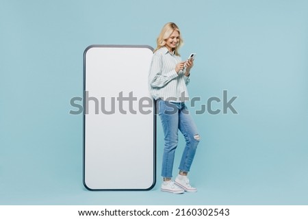 Full body elderly woman 50s in striped shirt near big white blank poster billboard for promotional content, place for text or image use mobile cell phone isolated on plain pastel light blue background