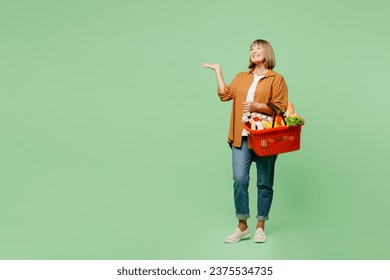 Full body elderly woman wear brown shirt casual clothes hold shopping paper bag with food products point hand arm aside on area mockup isolated on plain green background. Delivery service from shop - Powered by Shutterstock