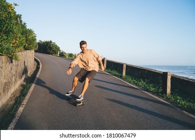 Full body of energetic man in casual clothes riding skateboard on empty embankment near sea and cloudless sky during sunny day - Powered by Shutterstock